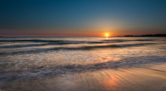  Die ersten Strahlen der gerade aufgegangenen Sonne brechen sich auf den Bewegungen der Wellen am Ufer des Timmendorfer Strands