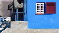  Blaue Hausfassade auf Graswader bei Heiligenhafen –  DETAIL: Das Foto zeigt eine blaue Hauswand mit einem weißen Fenster und rotem Laden. Das Haus steht – zum Schutz vor den Wellen und Hochwasser – auf einem Sockel aus Beton. Im Hintergrund erkennt man die typischen Details von Reetdach-Häusern.