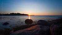  Ein sommerlicher Sonnenuntergang vor einer Steinbuhne bei Strande –  DETAIL: Die Sonne berührt den Horizont und taucht mit Ihrem Licht den Himmel in zartes Abendlicht. Im Gegenlicht zeigt das Foto am Horizont die Ausläufer der Eckernförder Bucht.